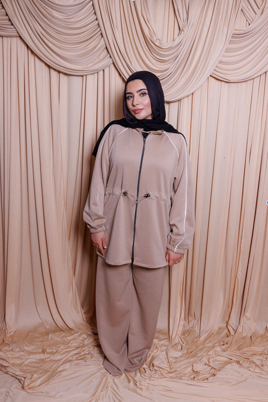 Woman wearing a beige outfit with a black hijab in front of beige curtains